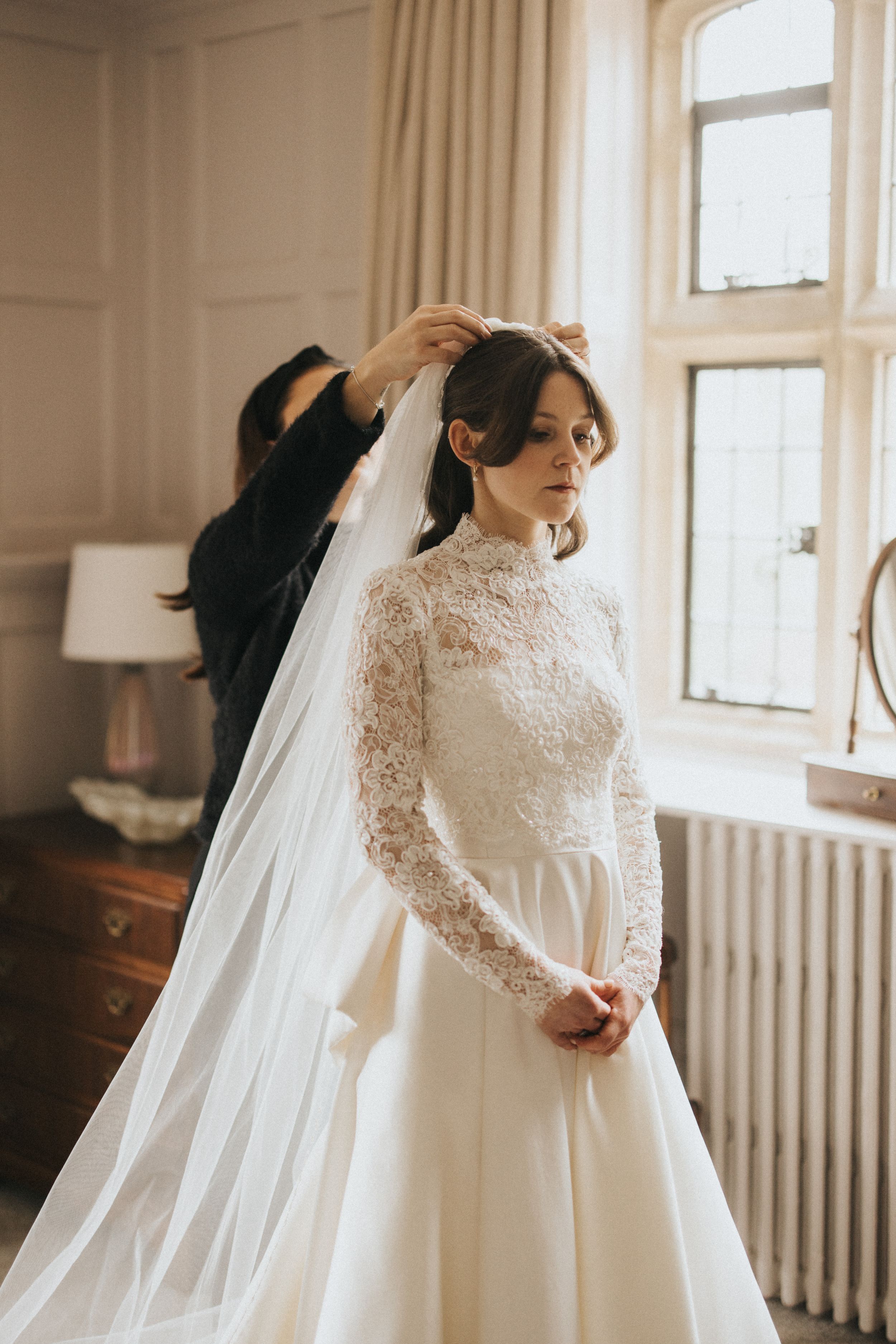 A bride wearing a long-sleeved lace wedding gown stands by a window while her veil is being adjusted.