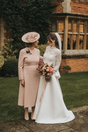 Wedding photography with figures in a vintage-style white bridal gown and blush pink suit against a brick mansion exterior.