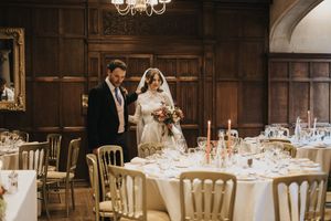 A couple stands together in an elegant wood-paneled dining room with round tables set for a formal dinner reception.