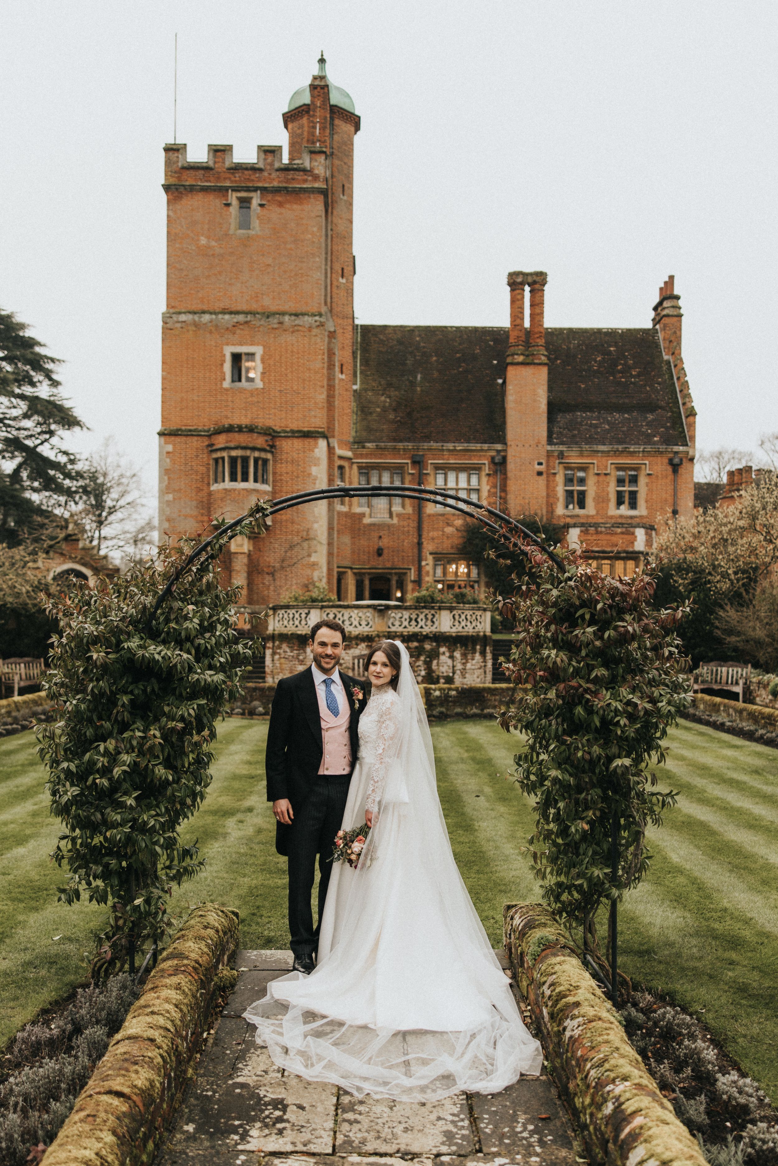 Two people stand on a garden path under a floral arch with a grand red brick Victorian tower building in the background.