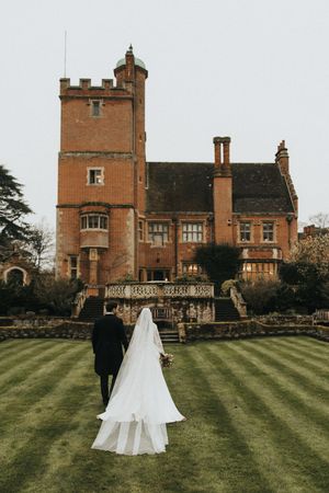 A romantic wedding couple strolls across the lawn in front of a majestic red brick Tudor-style castle mansion.