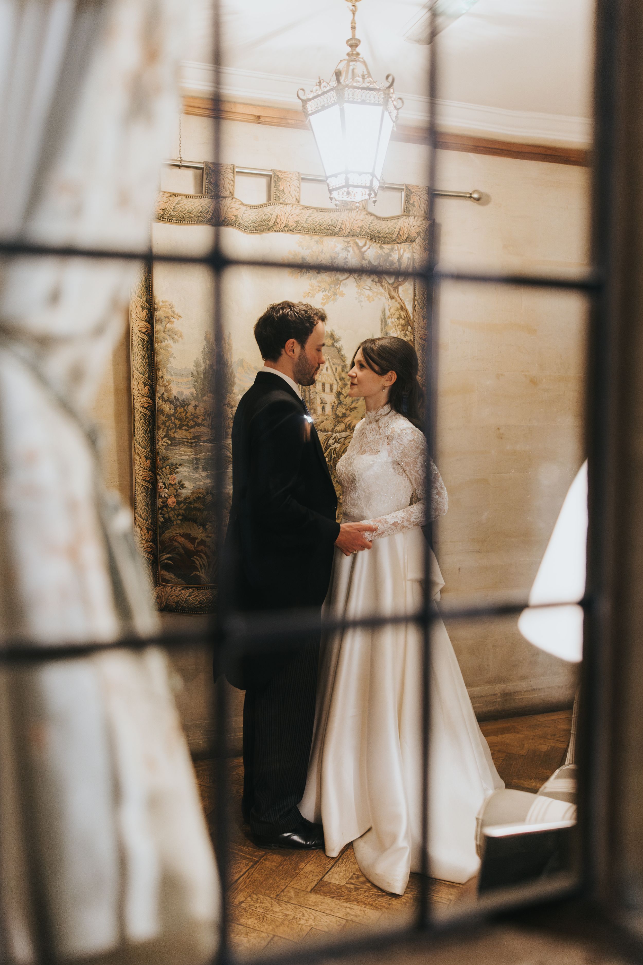 A romantic wedding moment captured through a window frame with an antique chandelier and mural in the elegant background.
