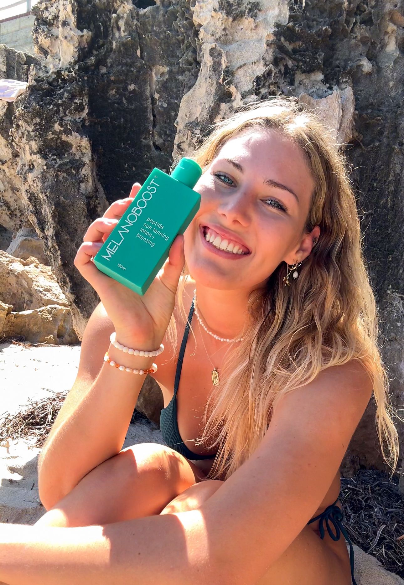Woman showcasing green beauty product bottle against rocky backdrop in sunny beach setting.
