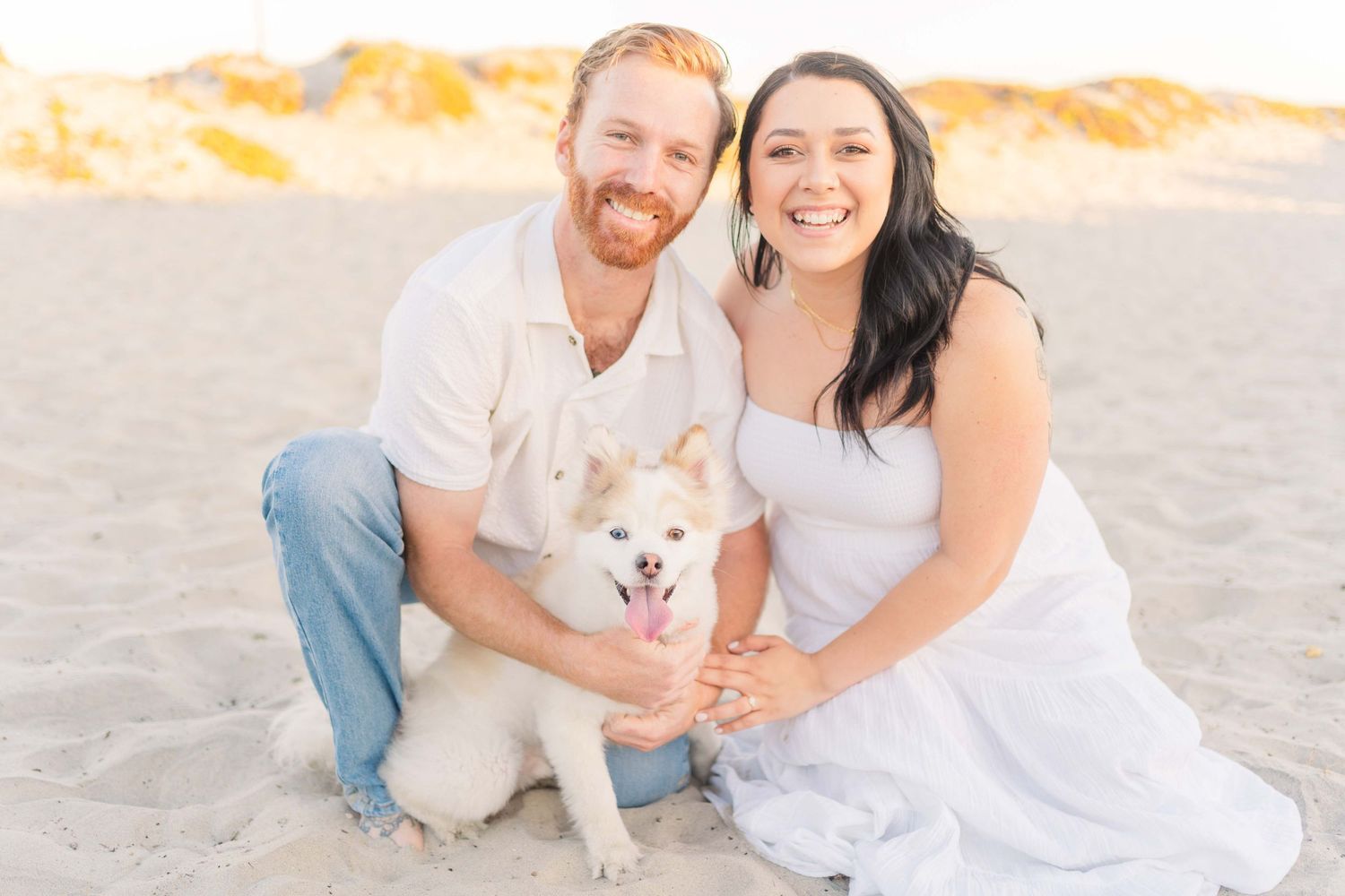 A couple in white clothes sits on a sandy beach at sunset with their small white dog.