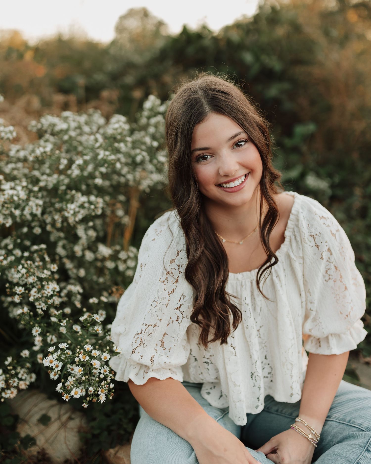 Smiling woman in a white lace top sitting outdoors against flowering white bushes and autumn foliage.