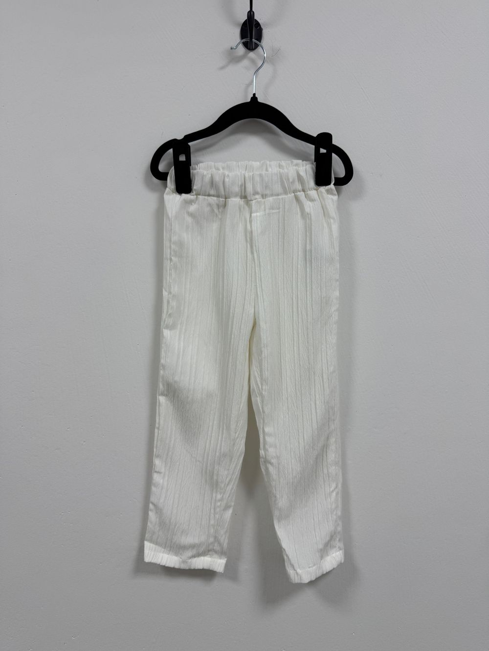 White linen pants with elastic waistband hanging on black clothes hanger against light gray background.