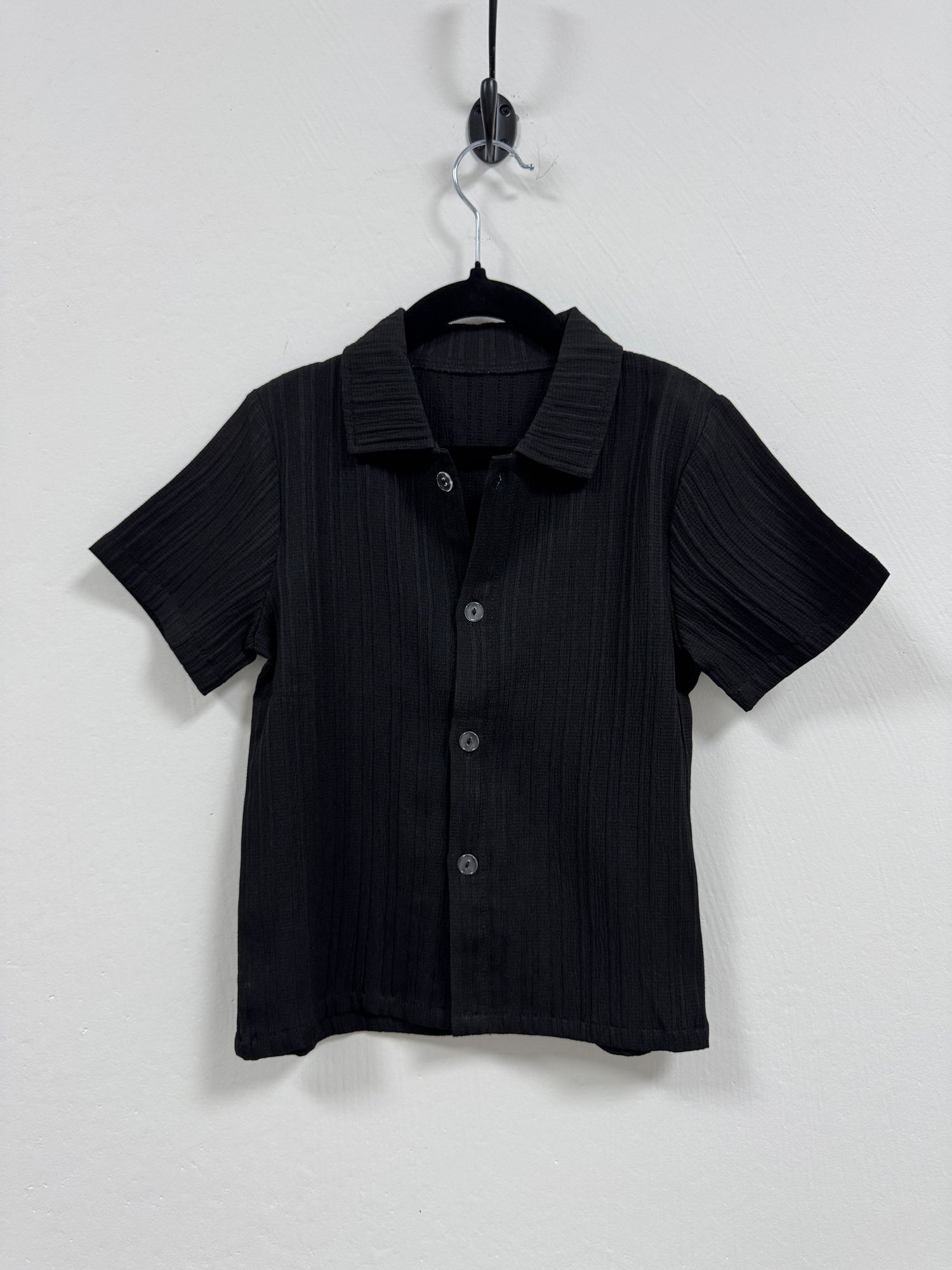 A black ribbed short-sleeve button-up shirt hanging on a clothes hanger against a white wall.