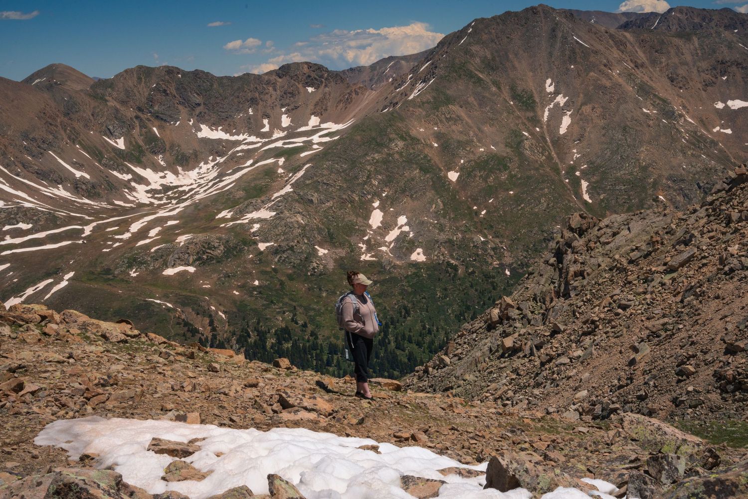 A hiker stands on snowy mountain terrain overlooking a dramatic alpine valley. Adventure session colorado