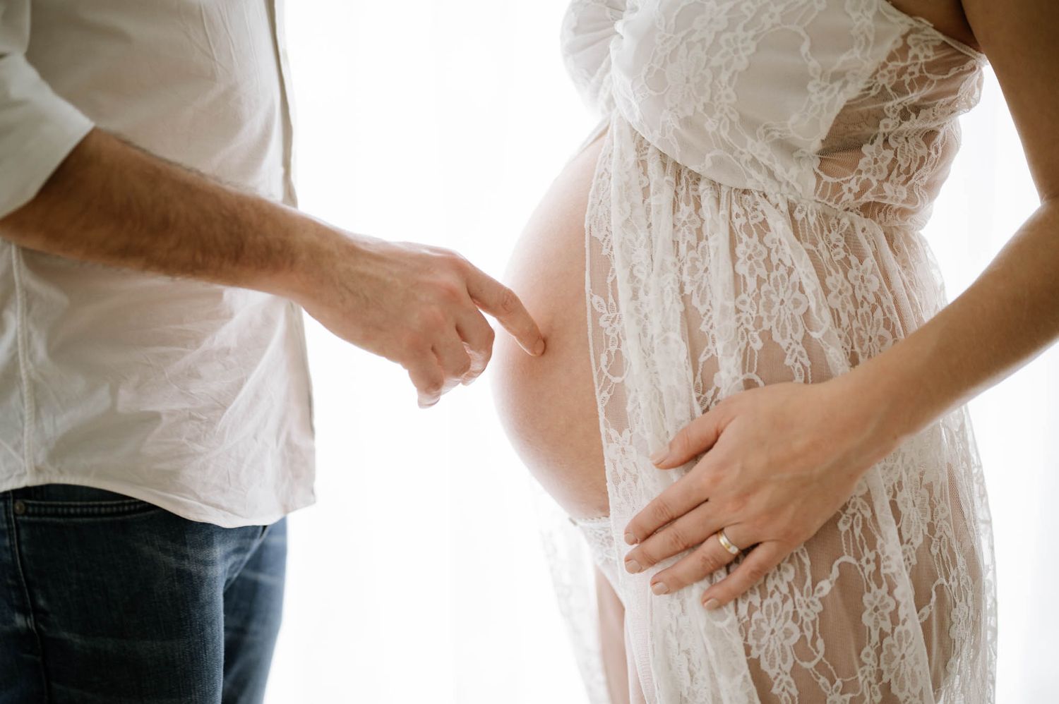 Close-up of pregnant belly with hands resting on it, wearing white lace dress and distressed jeans.