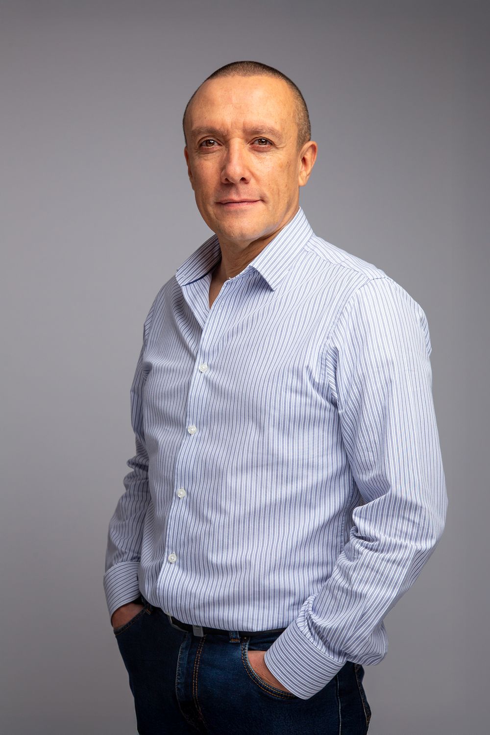 Professional studio portrait of a person wearing a light blue striped button-down shirt and jeans against a gray background.