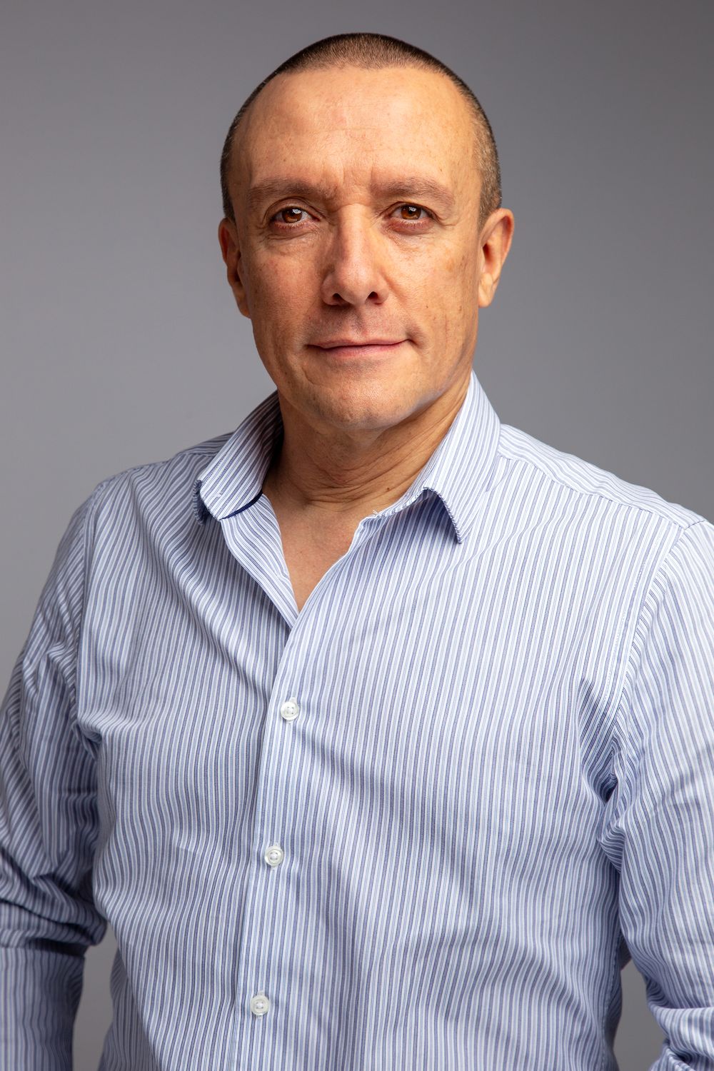 Professional studio portrait of a person wearing a light blue pinstriped button-down shirt against a gray background.