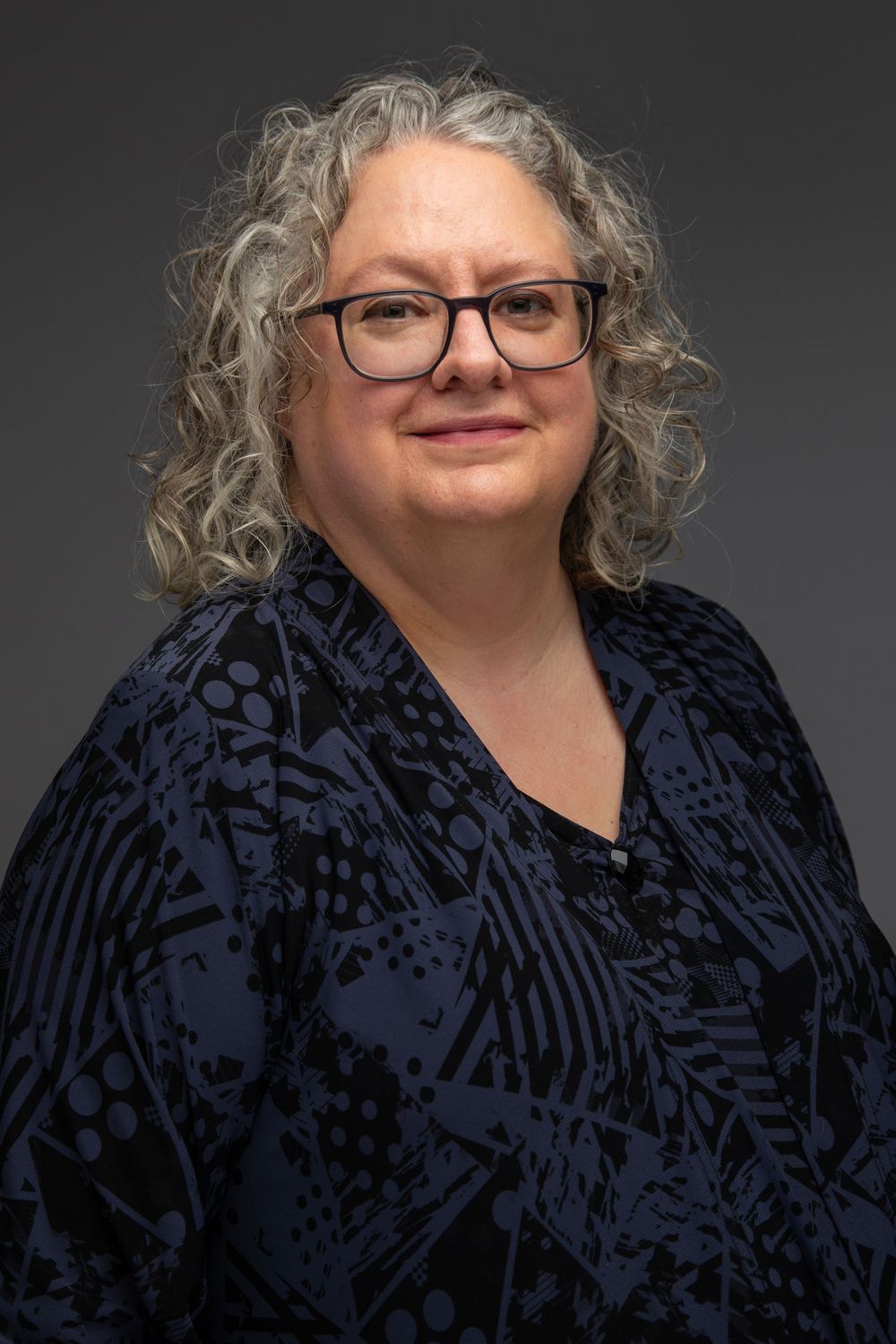 Professional headshot of someone with curly silver hair wearing glasses and a dark patterned top against a gray background.