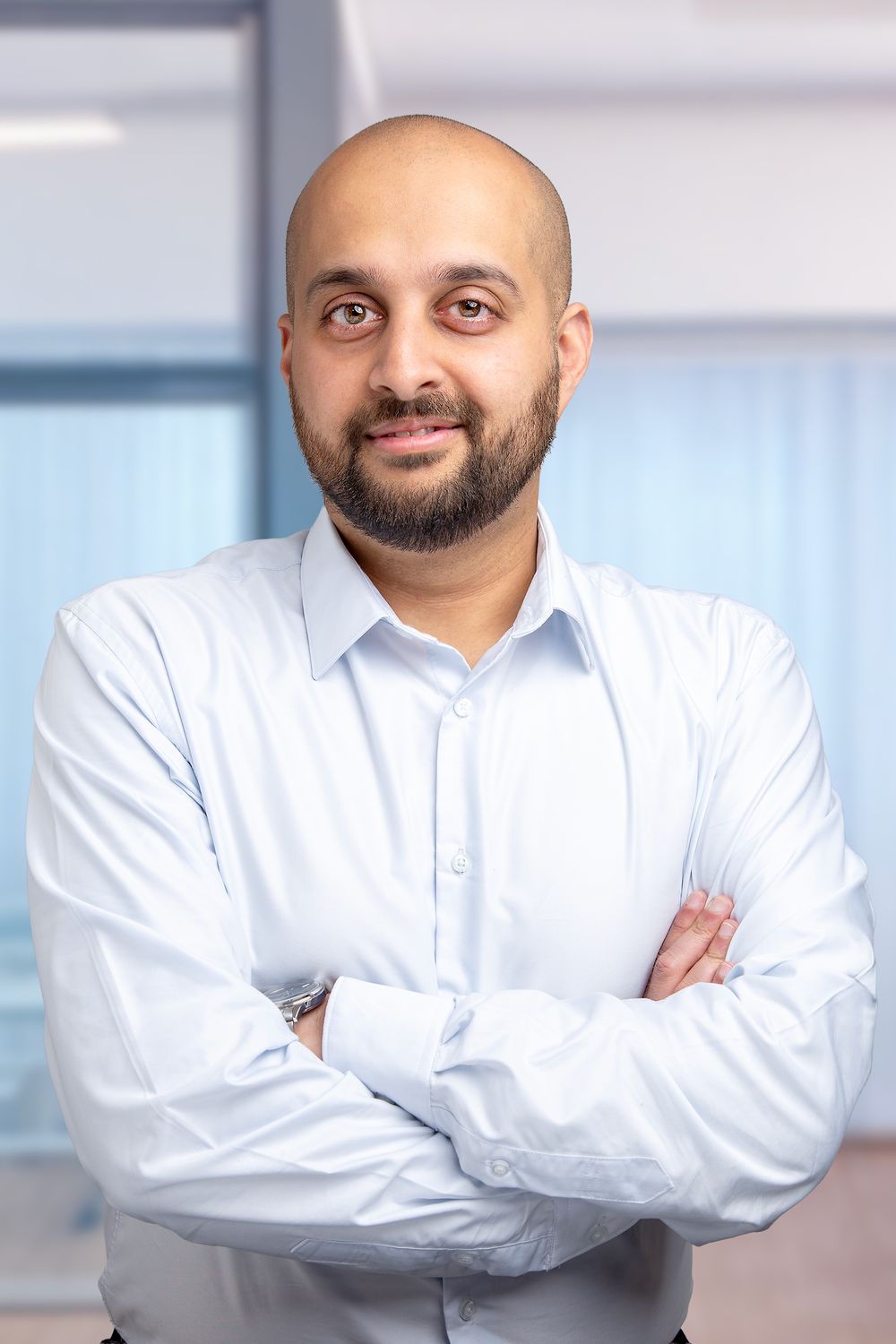A professional in a white dress shirt stands with arms crossed in an office setting with a confident expression.