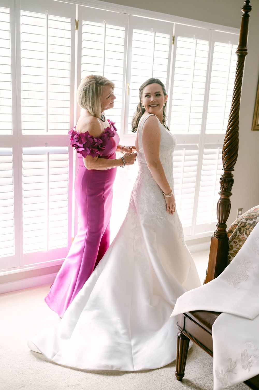 A wedding preparation scene showing a white gown and pink formal dress in a bright room with shutters.