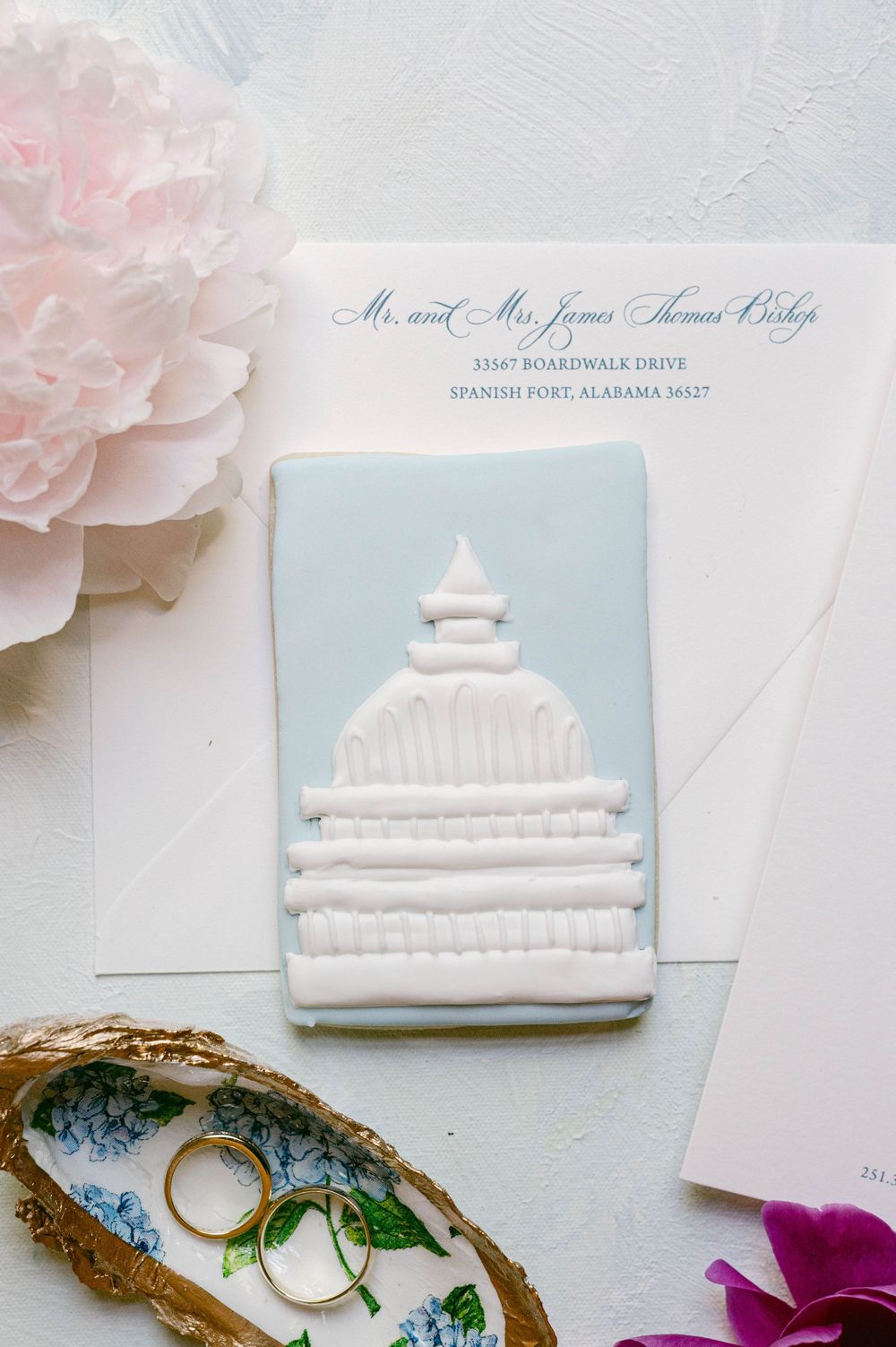 Wedding invitation with white architectural detail and rings beside pink peony flower.