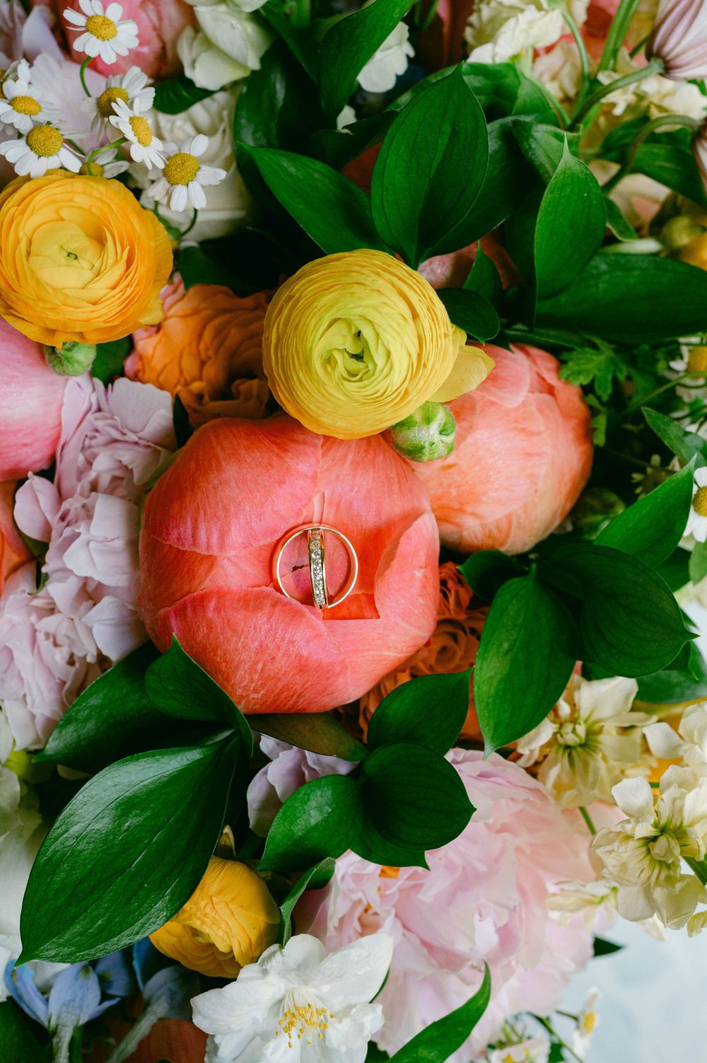 Wedding rings nestled in coral and yellow ranunculus flowers with green foliage.