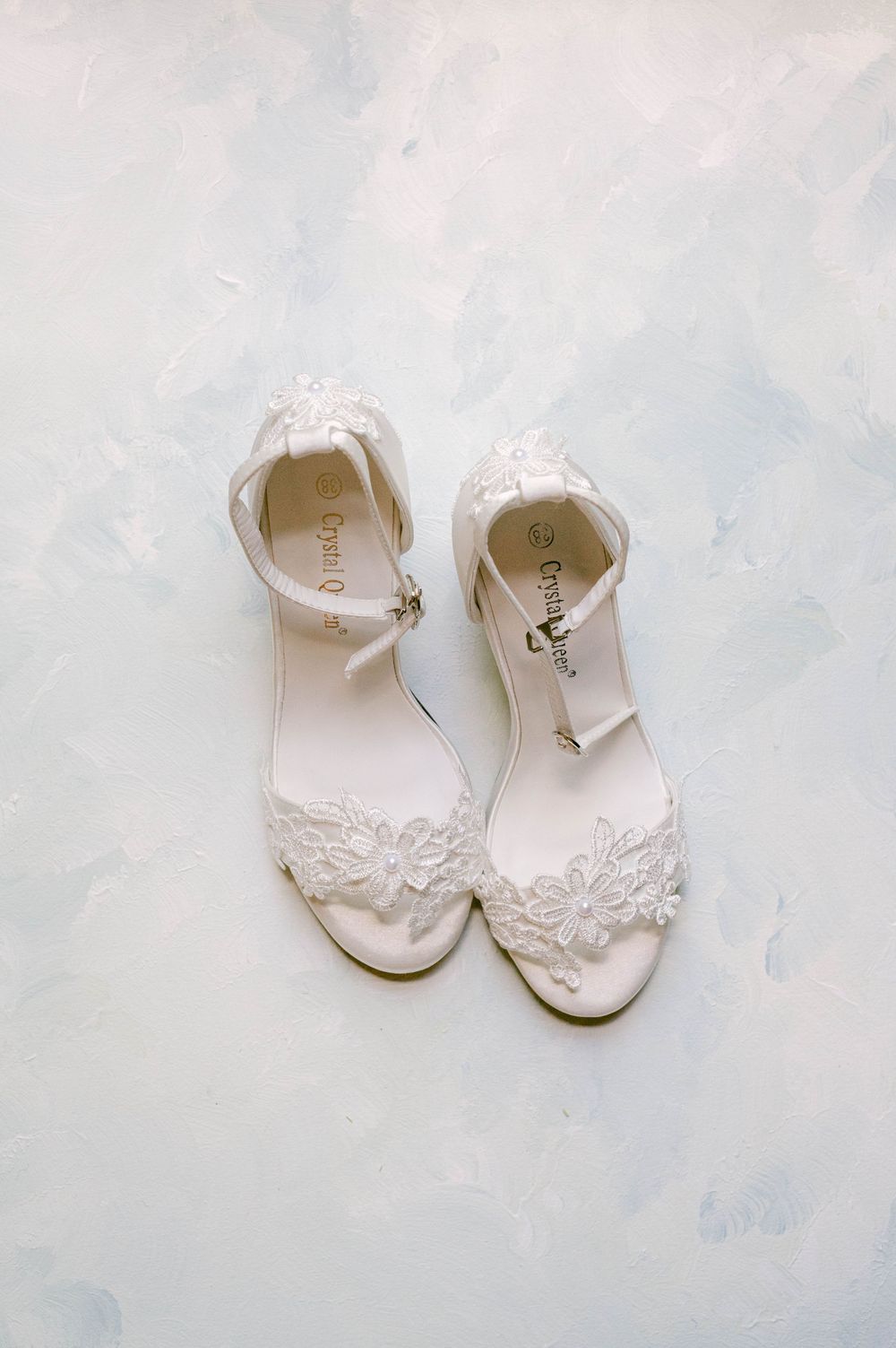 Delicate white bridal sandals with lace appliques and ankle straps on a light blue background.