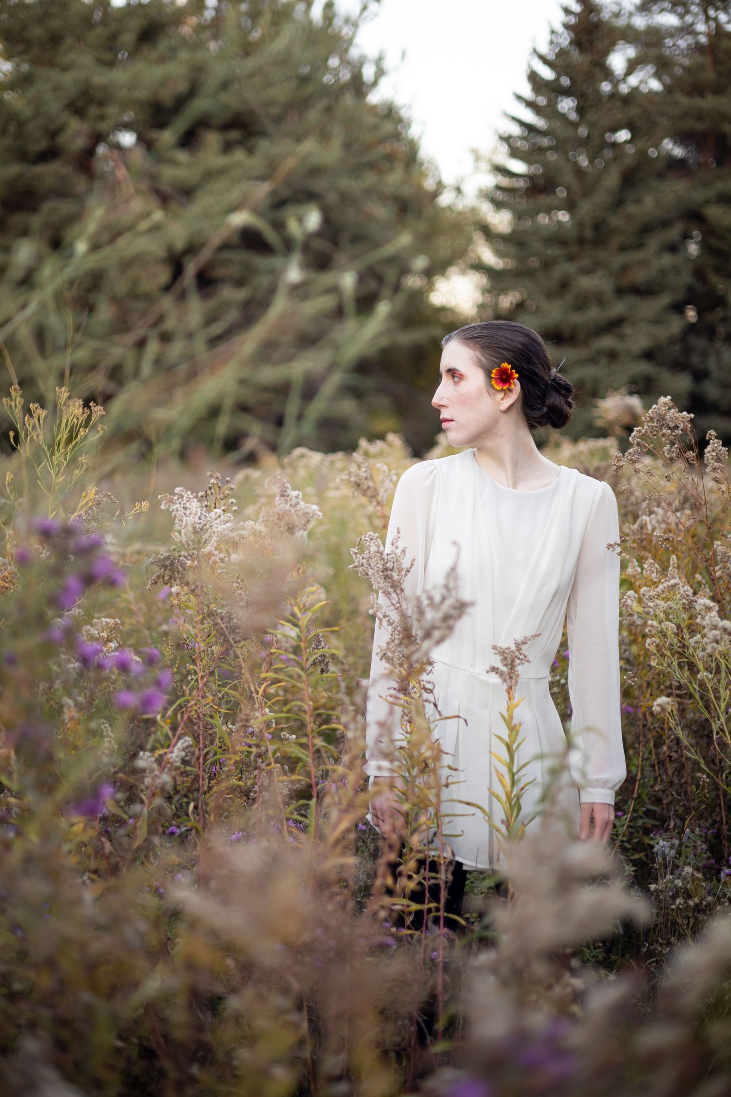 A person in a white blouse stands among wildflowers in a tranquil meadow with pine trees in the background during sunset.