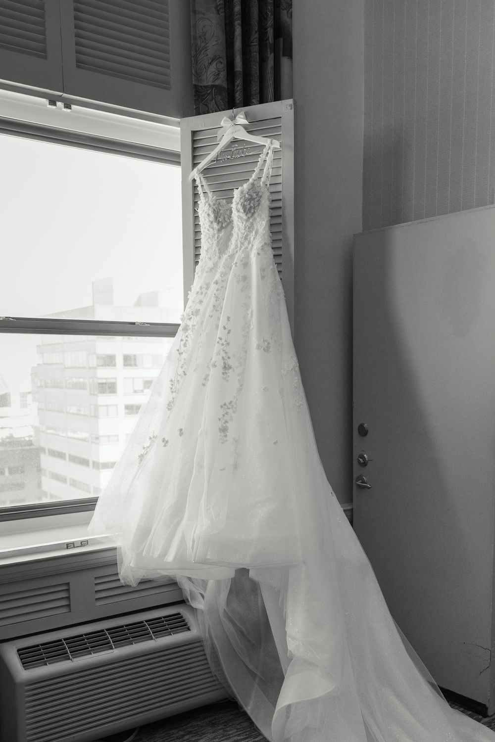 White wedding dresses hanging by windows in a hotel room preparation space.