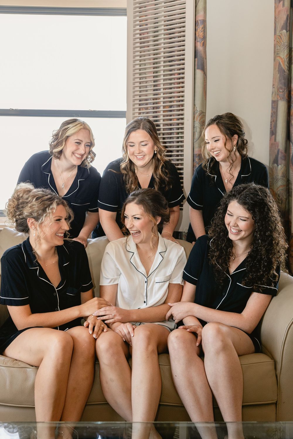Five bridesmaids in matching black pajamas sitting with the bride in white on a beige couch.