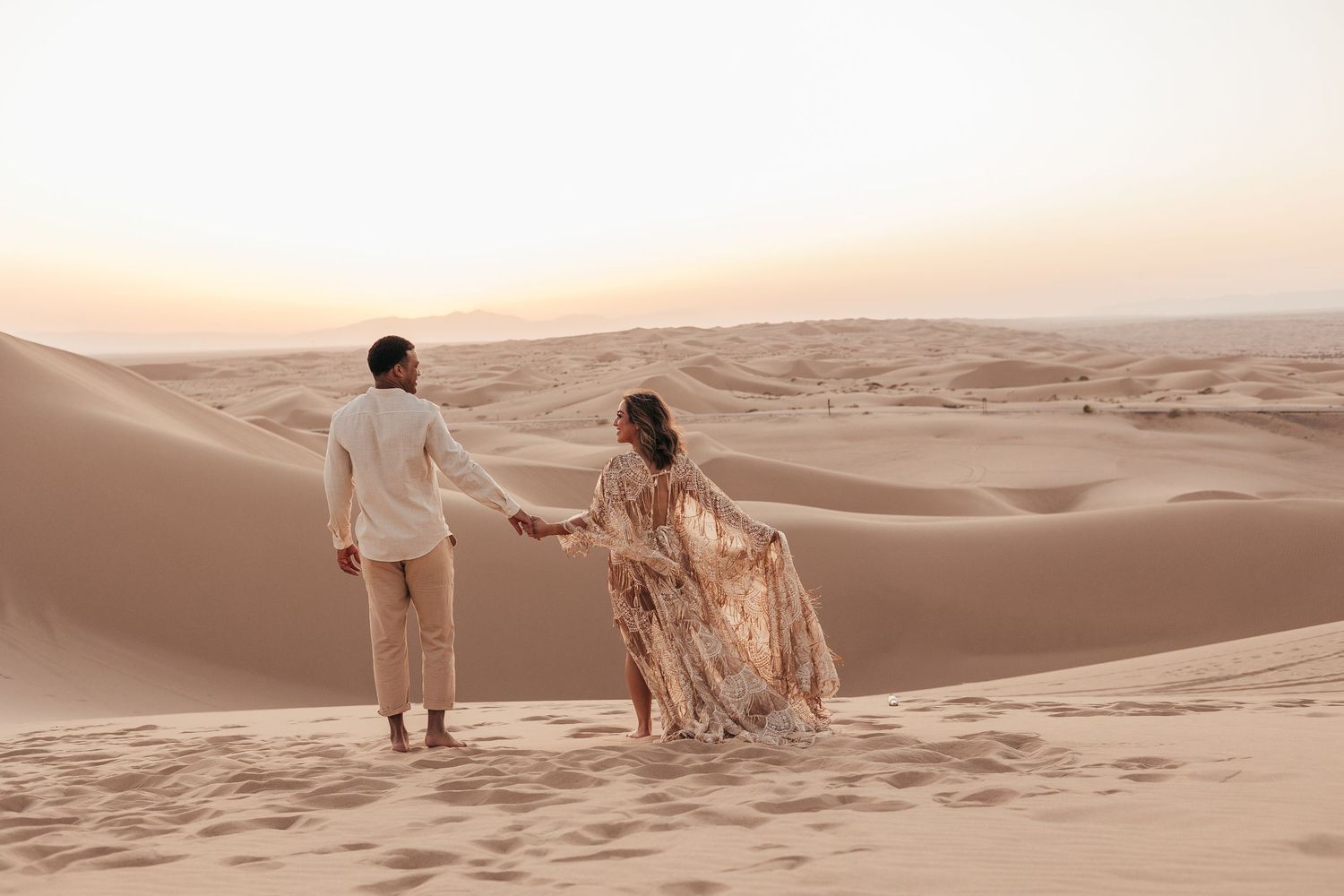 Two people holding hands while standing on sand dunes during sunset in a desert landscape.