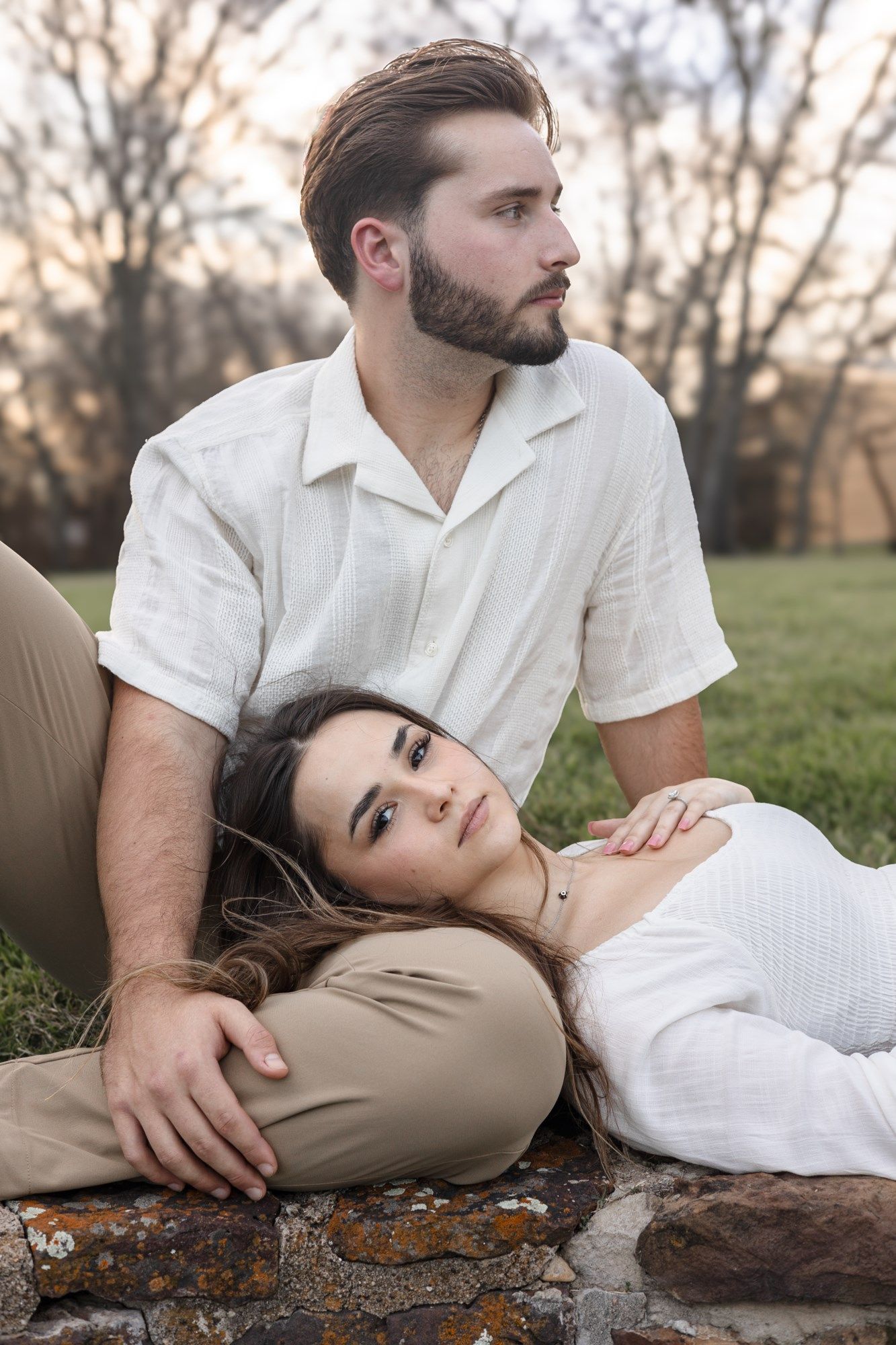 A romantic couple in white clothing lounges together on a stone wall at sunset in a grassy outdoor setting.