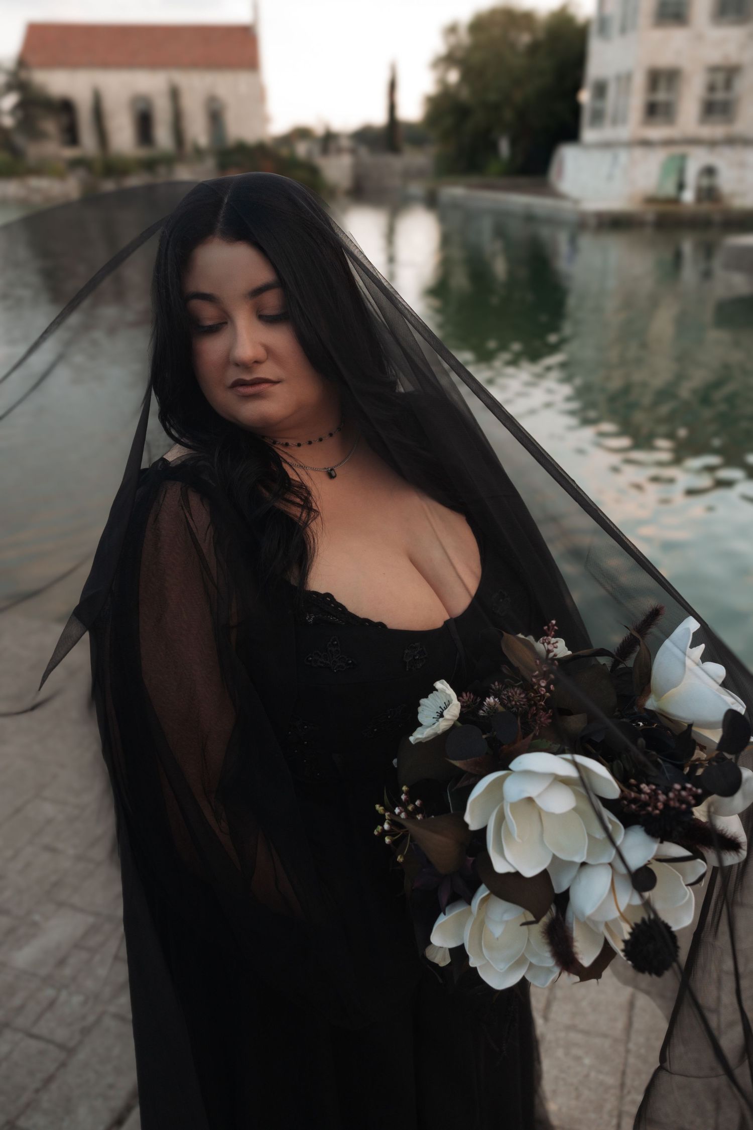A figure in a black veil poses near water with white orchids, creating a moody gothic aesthetic.