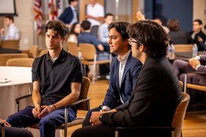 Three people sit on chairs during a panel discussion at an event with an American flag visible in the background.
