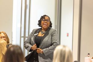 A woman in a gray blazer speaks enthusiastically to an audience at a professional event or conference.