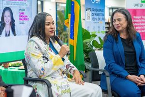 Two women speak at a Brazil-themed event, one holding a microphone in a floral outfit, the other in a blue blazer.
