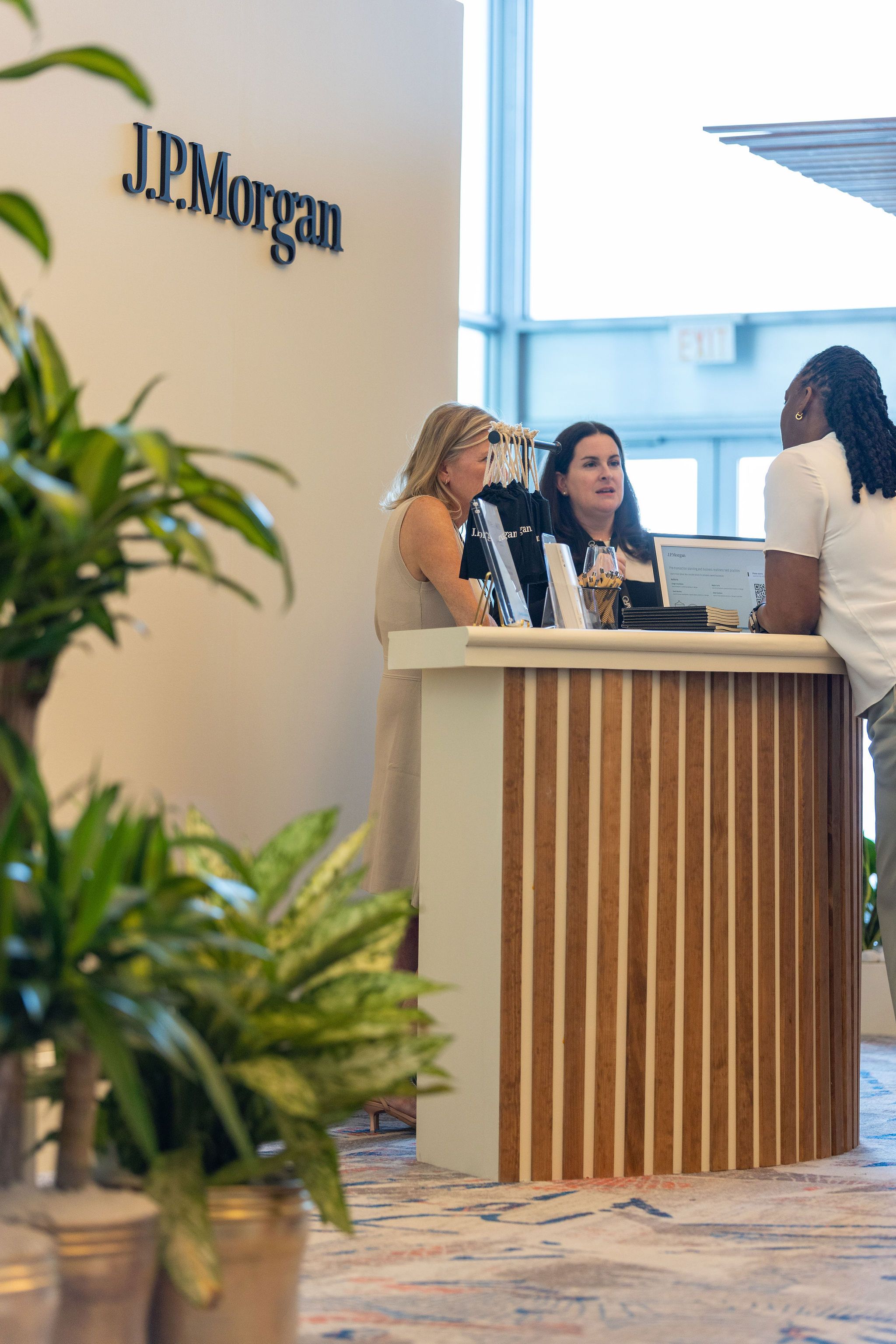 Two women working at a modern reception desk beneath a J.P. Morgan sign with green plants in foreground.
