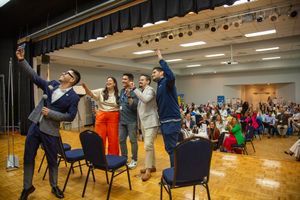 Performers on chairs entertain a crowd during an interactive stage show in a school gymnasium.