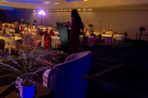 A speaker presents at a dimly lit conference hall with round tables of attendees, a blue chair and palm plant in the foreground.