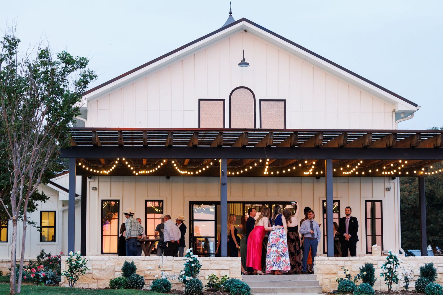 Evening wedding celebration at Bella Cavalli,a white farmhouse venue, with string lights illuminating the covered porch where guests gather.