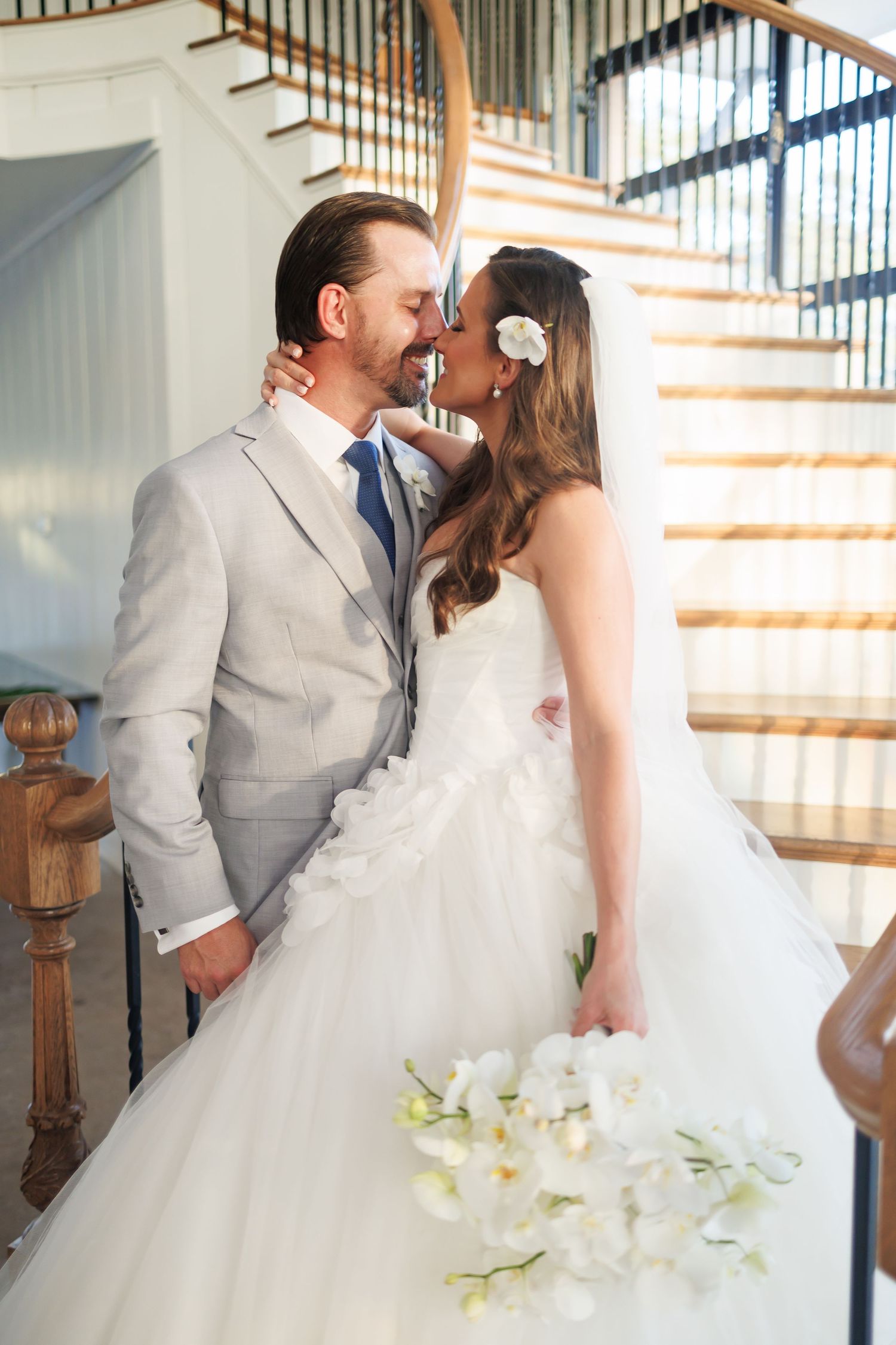 Couple kisses on elegant curved staircase at Bella Cavalli with large windows, bride in flowing white gown holds white floral bouquet.