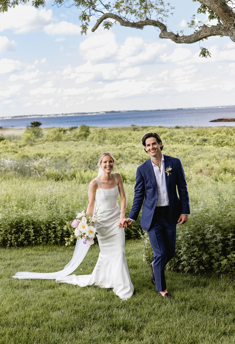 Couple walks through a lush green field with water views in the background. Nonquitt Beach. New England Wedding.