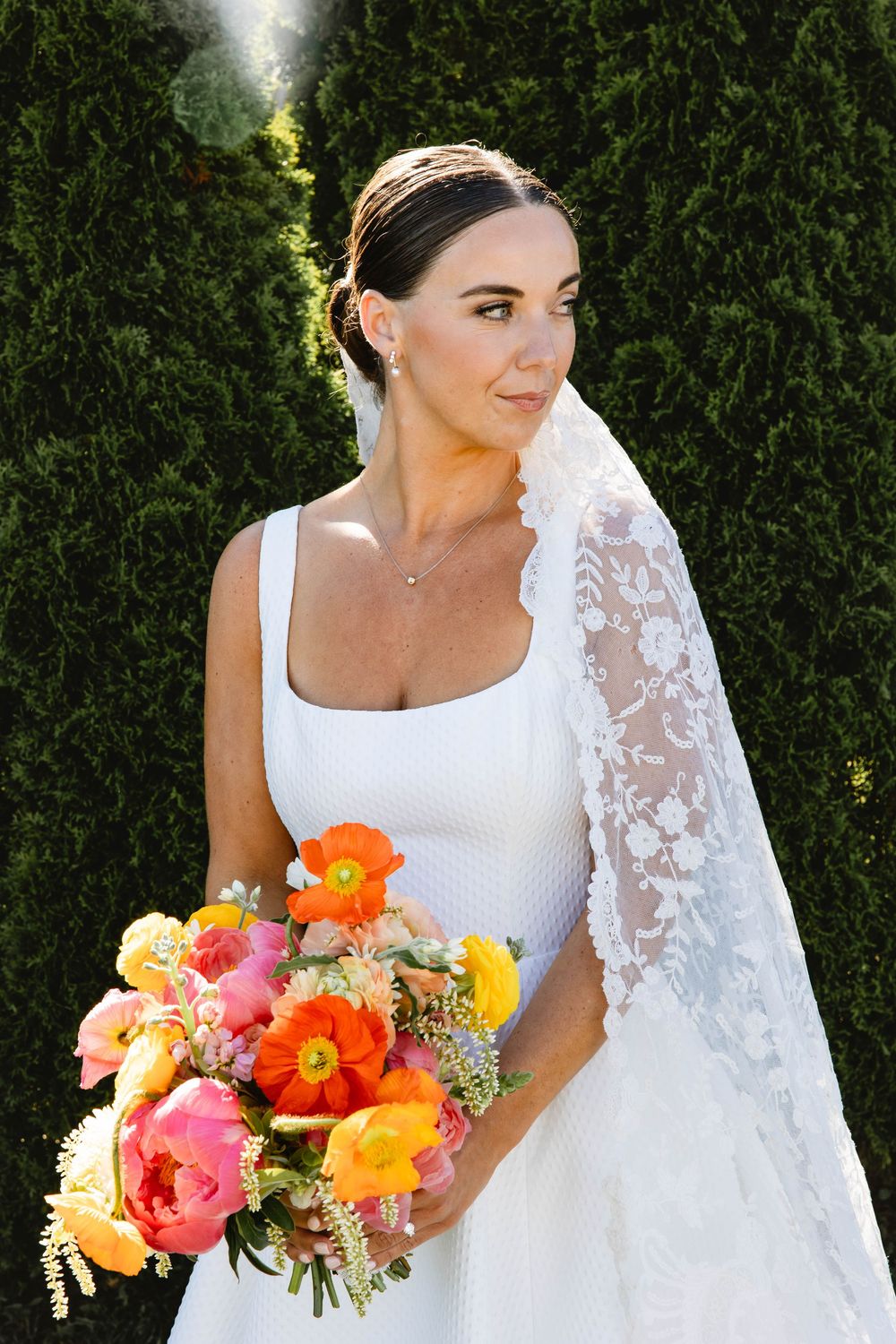 Bride in white dress holding vibrant orange and pink floral bouquet against green hedge backdrop.