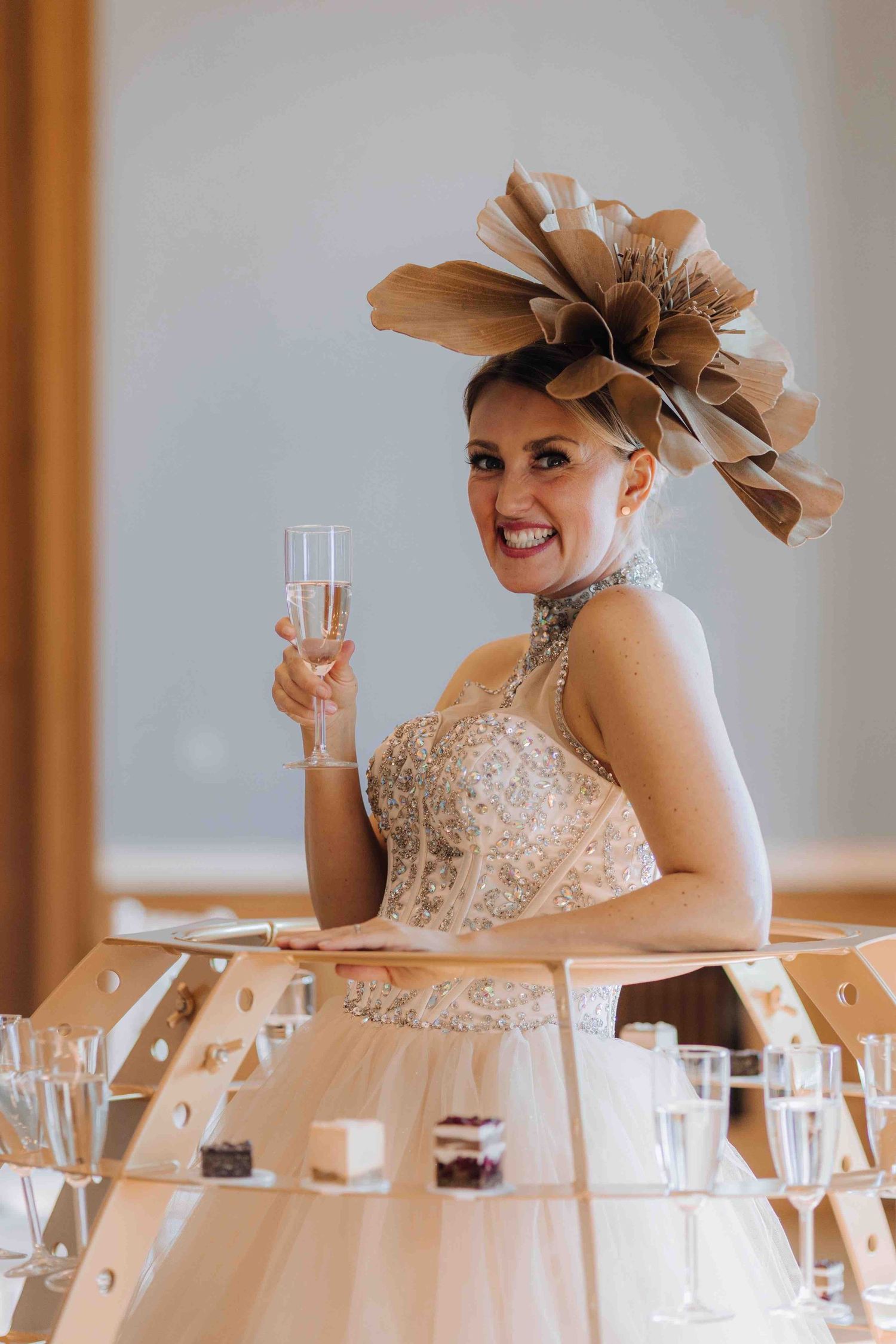A bride in a champagne dress and dramatic brown floral headpiece holds up a glass beside a champagne tower display.