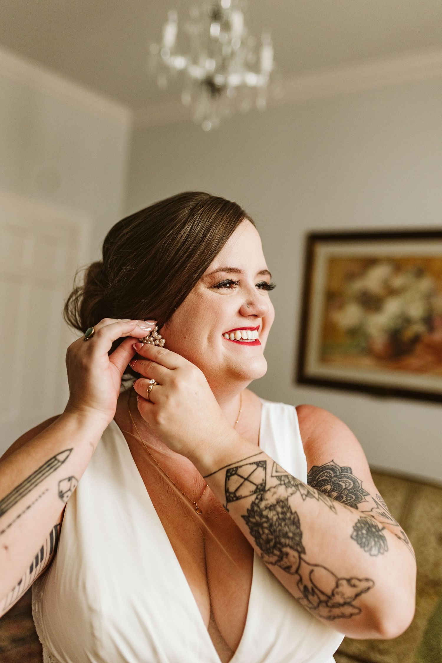 A person in a white wedding dress smiles while putting on jewelry, showing tattooed arms.