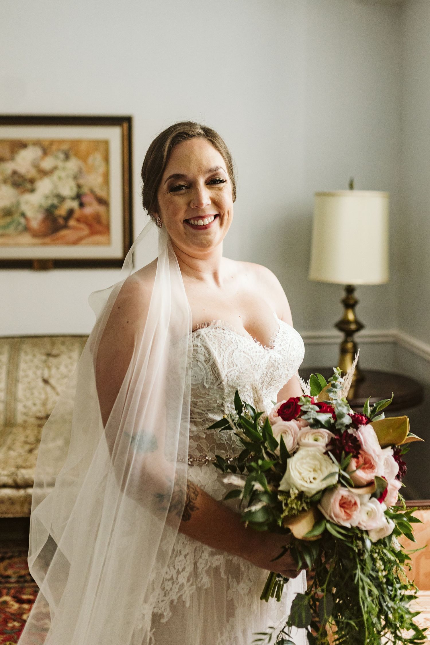 A bride in a lace wedding dress holds a colorful bouquet of flowers against a light interior wall.