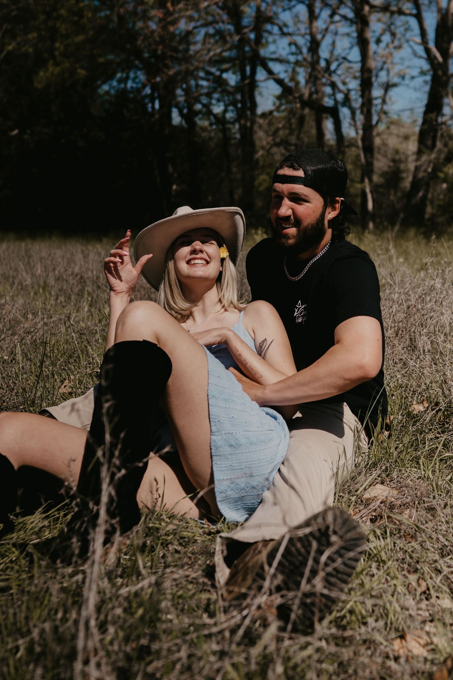 A couple shares a joyful moment sitting together in a sun-dappled meadow with trees in the background.