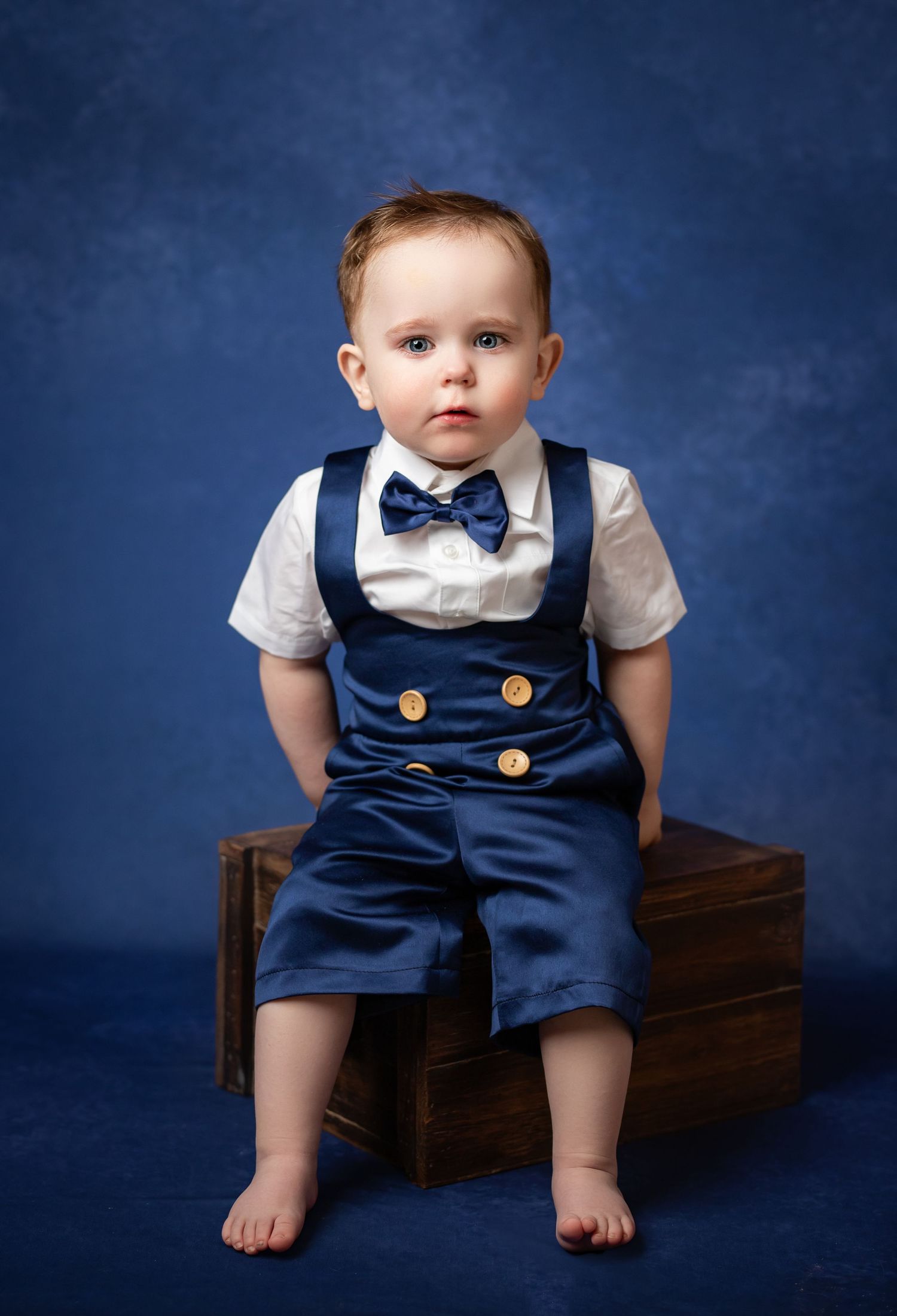 A small child in a formal navy blue overalls outfit with bowtie sits on a wooden box against a dark blue backdrop.