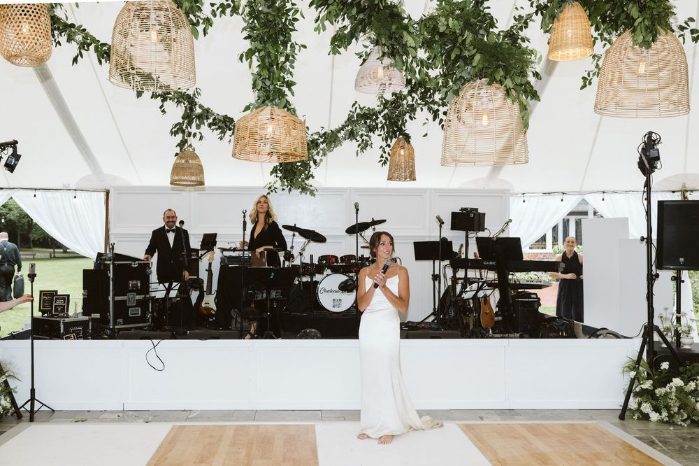 Bride in white gown speaks at wedding reception under wicker pendant lights with live band performing on stage.
