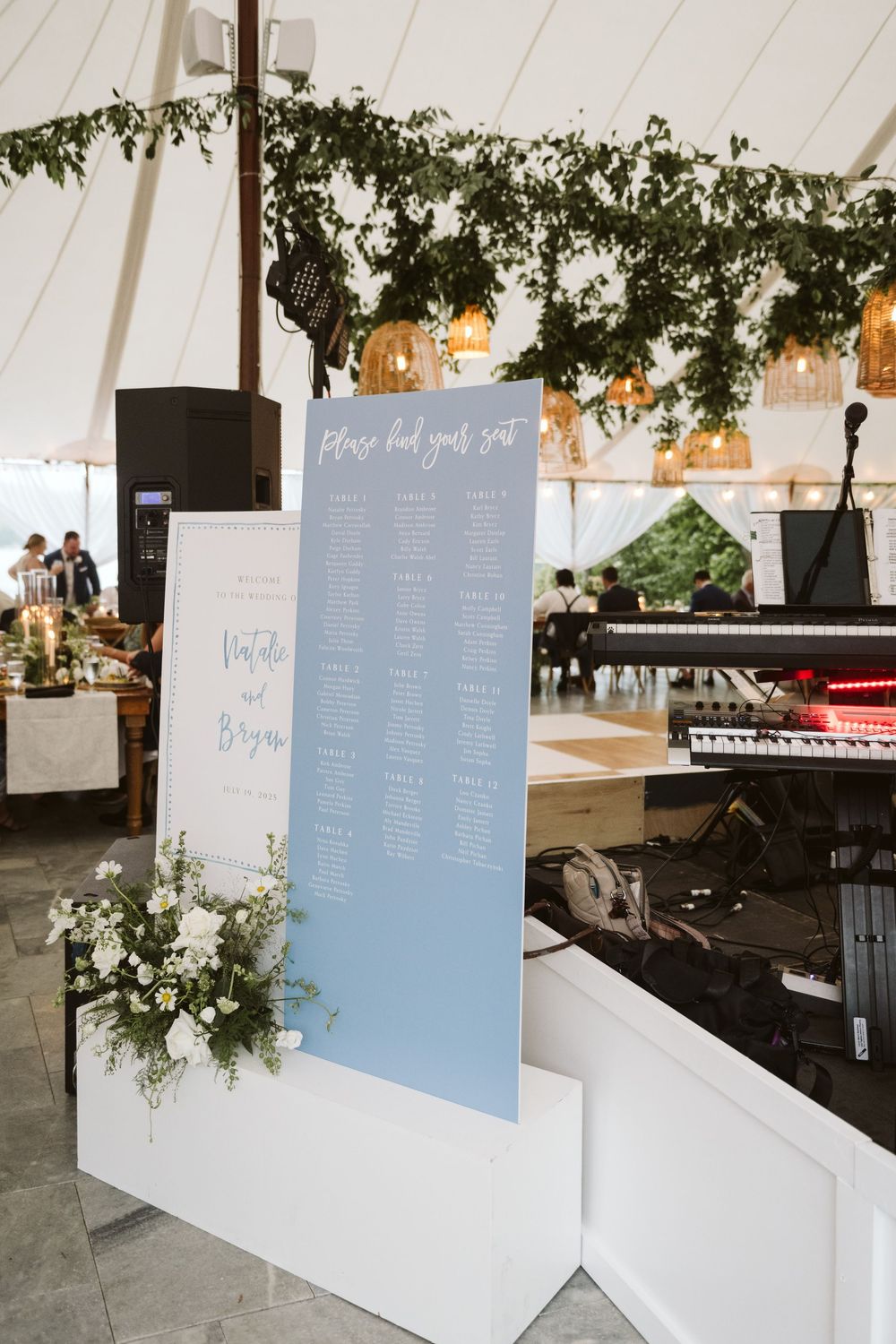 Elegant wedding seating chart display on dusty blue board with white floral decor inside a tented reception venue.