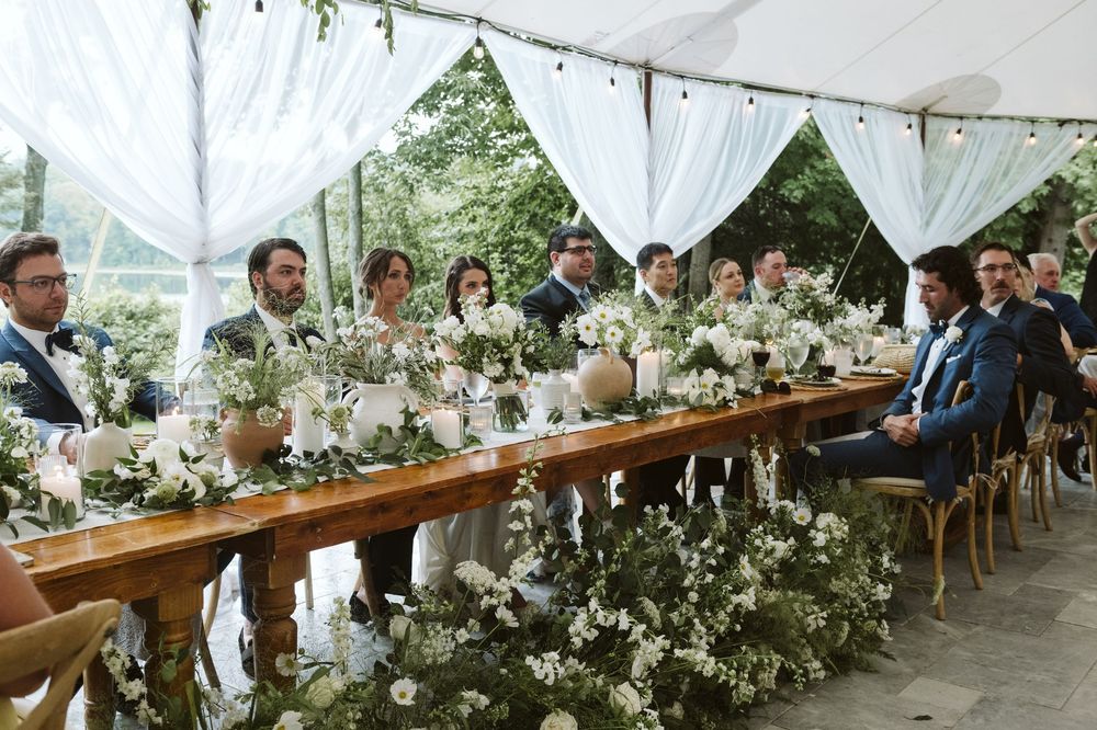 Wedding guests seated at a long decorated table adorned with white floral arrangements under a draped tent.