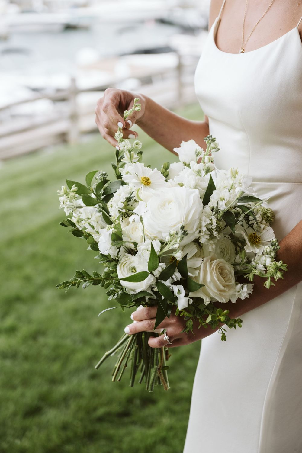 Bride in white gown holding lush white floral bouquet with roses and greenery outdoors on green lawn.