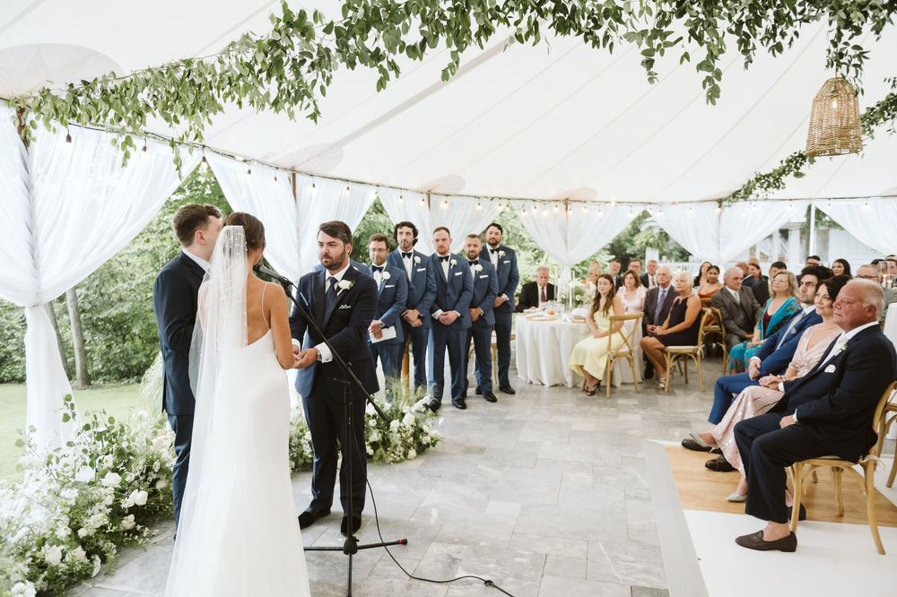 Bride and groom exchange vows under elegant white tent with floral greenery, groomsmen and seated guests in background.