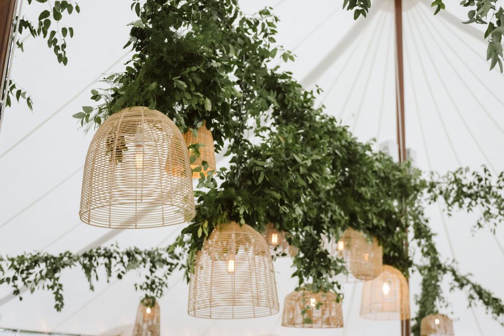 Rustic wicker pendant lights hanging from a white tent ceiling adorned with lush green garlands at a wedding reception.