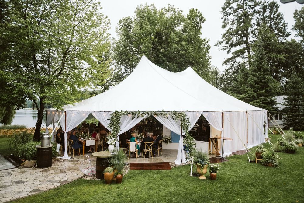 Elegant white sailcloth wedding tent set up outdoors on green lawn surrounded by lush trees and garden decor.