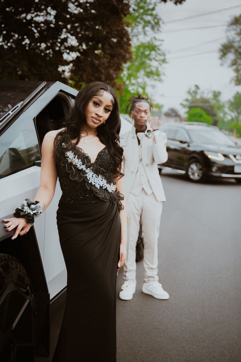 A young couple dressed in prom attire poses near a white limo, she in a black gown and he in a cream suit.